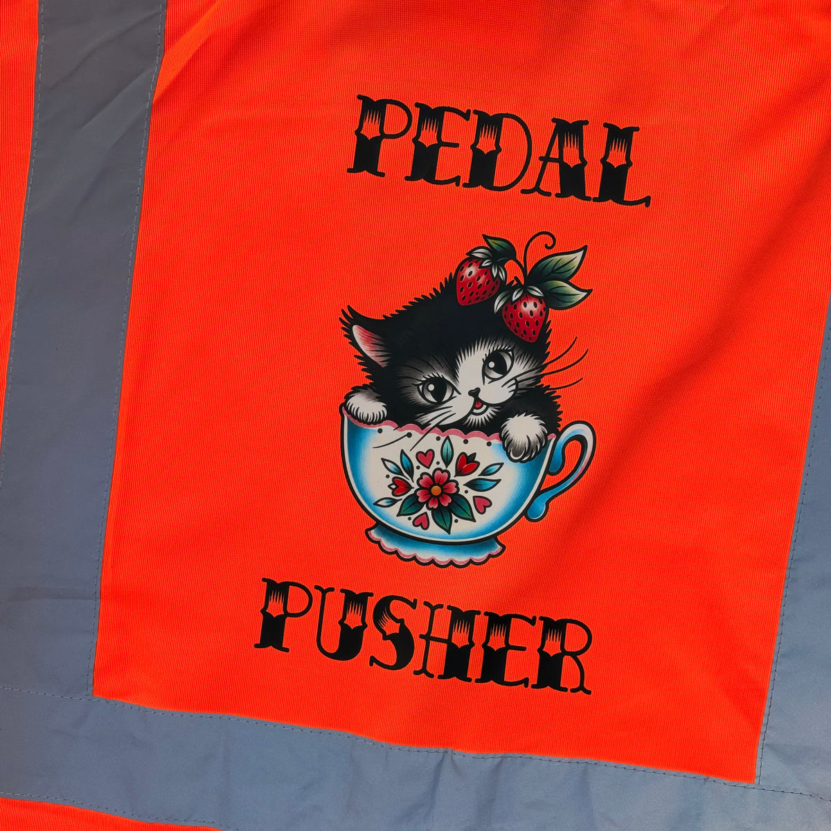 Close-up of a bright orange high vis cycling vest with a central graphic showing a black-and-white kitten in a floral teacup with strawberries, surrounded by the words “PEDAL PUSHER” in black.