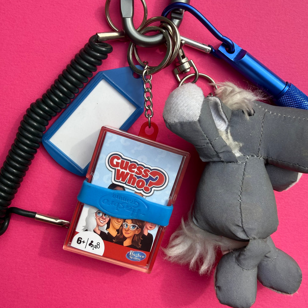 A plush gray sheep keychain with white wool details, attached to a metal keyring alongside a blue Guess Who game box, a black coiled cord, a blue carabiner, and a blank blue ID badge holder, all displayed on a bright pink background.