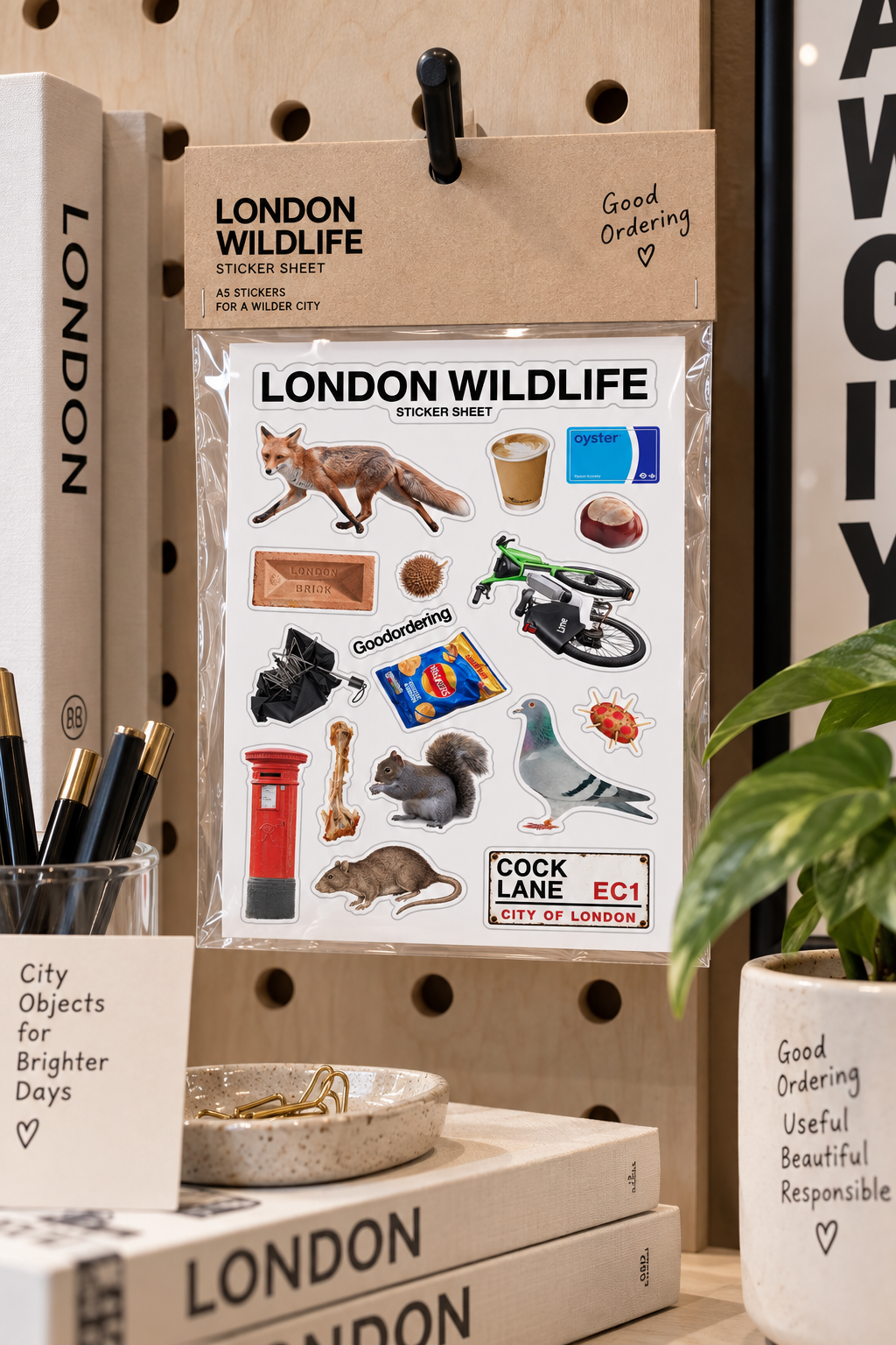 A packaged London Wildlife sticker sheet hanging on a pegboard shelf, surrounded by books, pens, and a plant. The sticker sheet features illustrations of a fox, pigeon, mouse, postman, red telephone booth, bicycle, and other London-themed motifs with text including 'LONDON WILDLIFE' and 'COCK LANE EC1 CITY OF LONDON'.