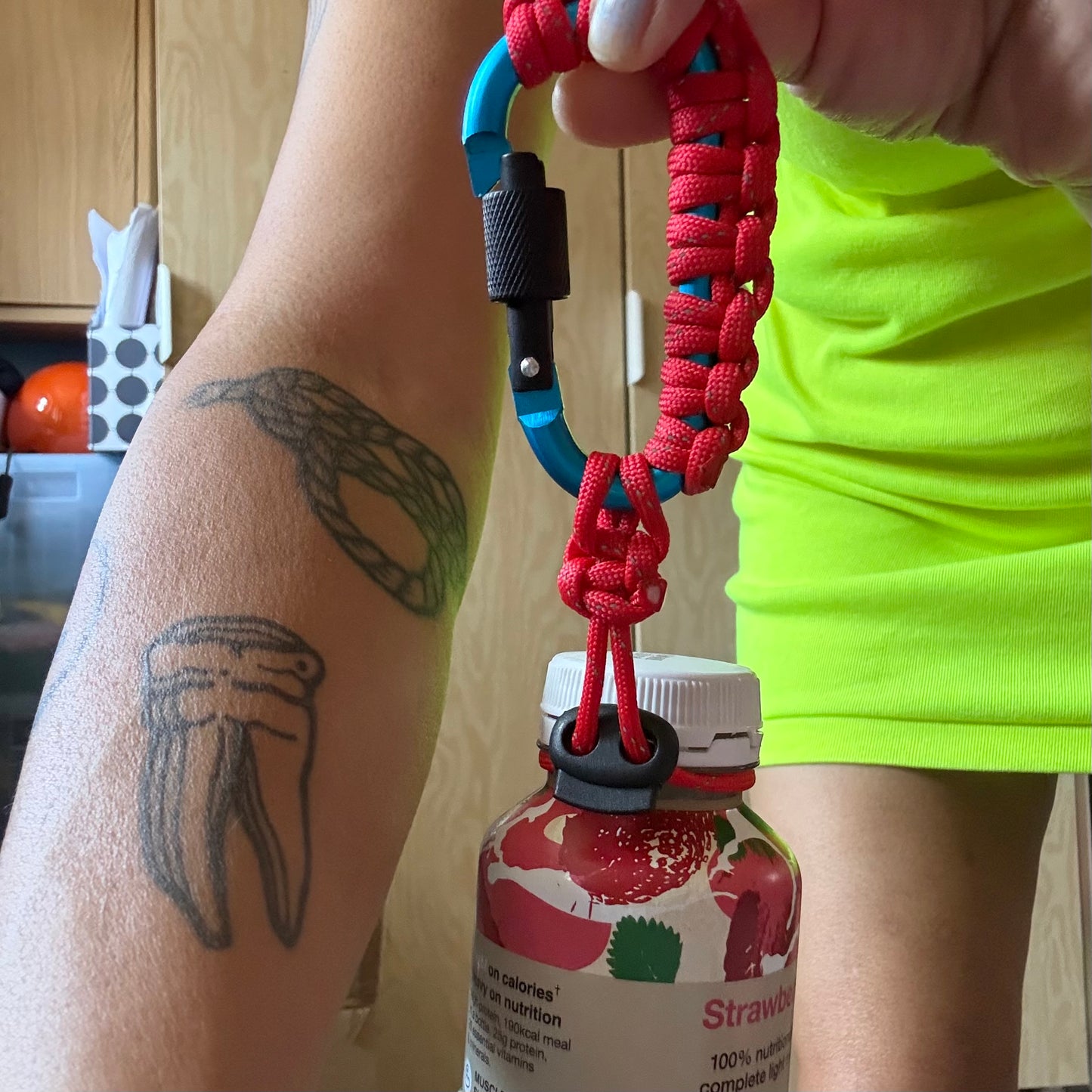 A close-up lifestyle shot of a red paracord bottle strap attached to a blue carabiner and held by a person's hand, with a person's tattooed forearm and bright neon yellow shirt visible in the background.