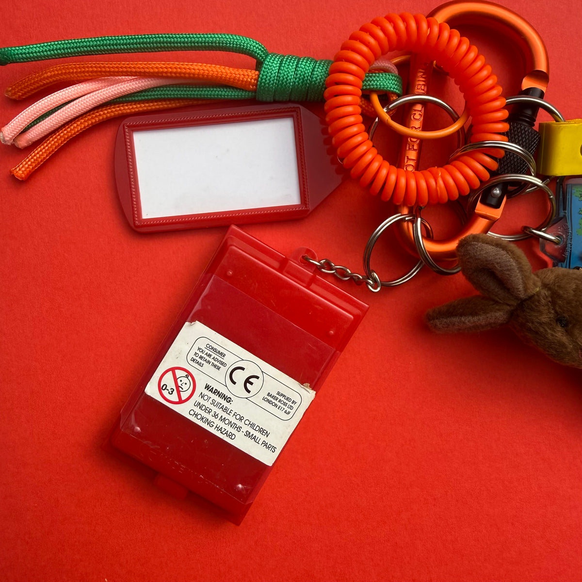 A red plastic personal safety keychain set on a red background, including a blank red ID tag, a coiled orange safety cord, a red alarm battery holder with a warning label, and small accessories.