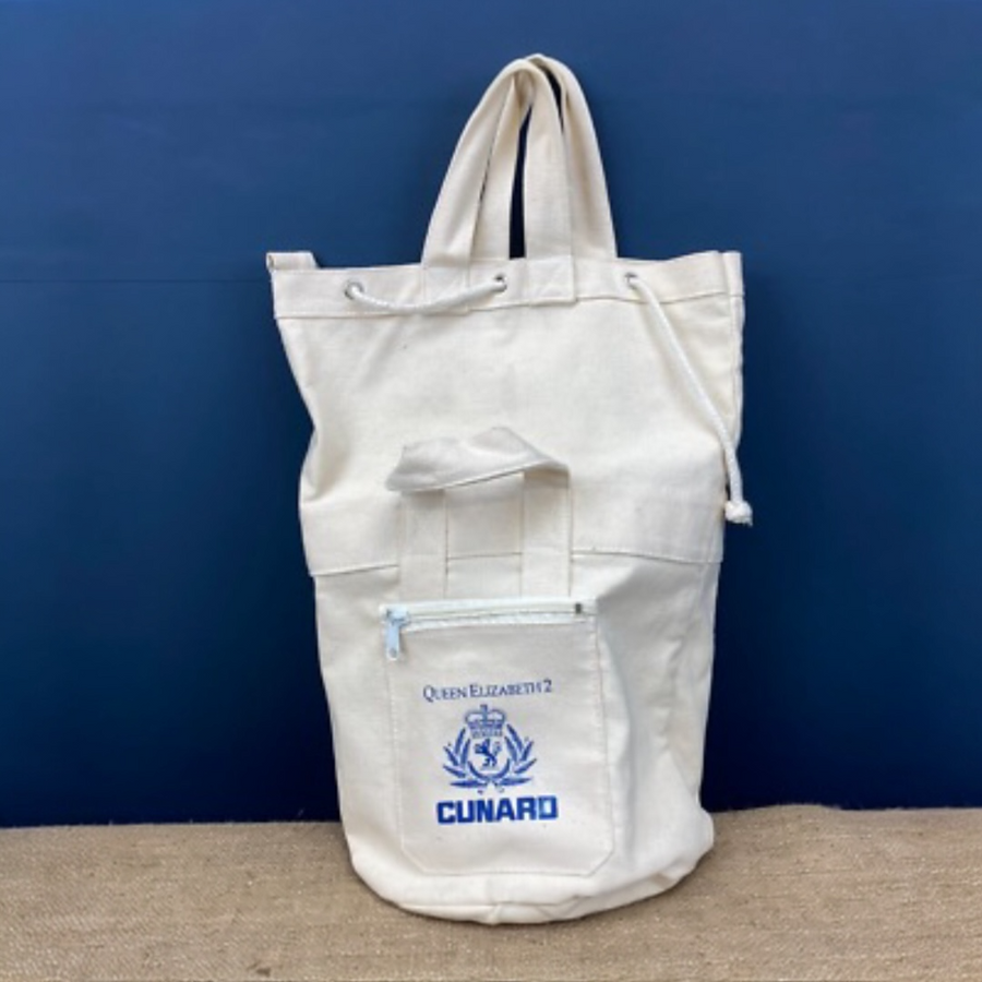 An off-white canvas tote duffle
bag with a front zippered pocket featuring a blue printed emblem and the text 'QUEEN ELIZABETH II CUNARD', set against a dark blue background.