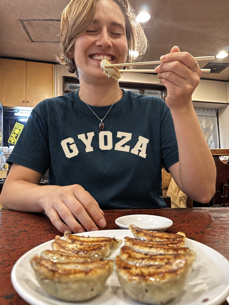 A person sitting at a restaurant table wearing a navy blue t-shirt with 'GYOZA' printed in white across the chest, holding chopsticks with food above a plate of pan-fried gyoza dumplings.