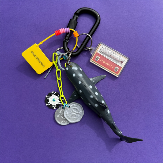 A shark-shaped keychain with a black carabiner, yellow and neon chain, coin charms, a small card, and a yellow tag, displayed on a purple background.