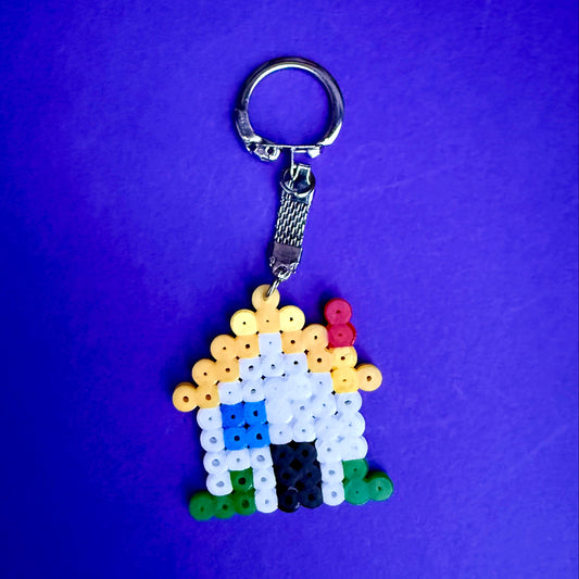 A handmade rainbow house keychain made of small colored beads, attached to a silver metal split ring and clasp, set against a solid purple background.