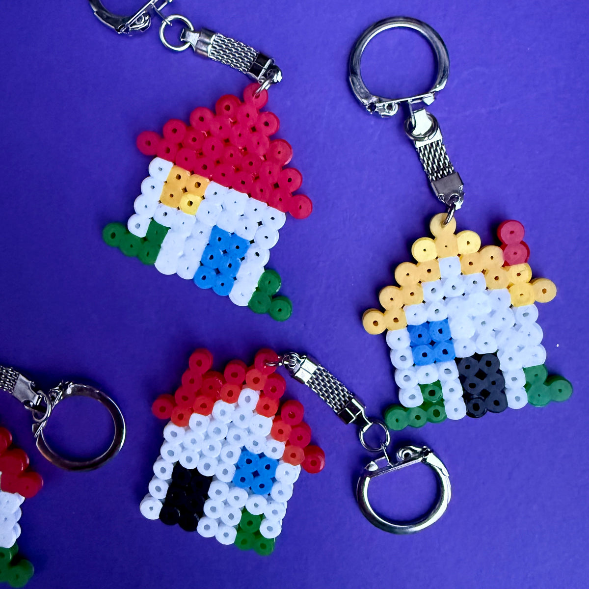 Several handmade Hama beads keychains shaped like houses are arranged on a solid purple background. The keychains feature silver metal keyrings and clasps, with houses crafted from white, red, blue, green, yellow, and black beads.