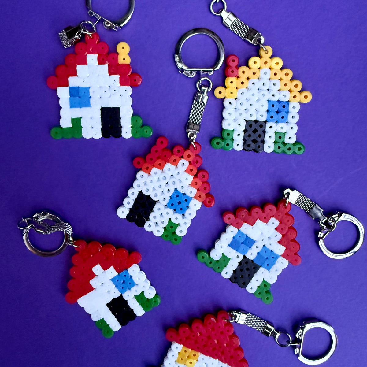 Six handmade house-shaped keychains crafted from colorful fidget beads, arranged on a solid purple background.