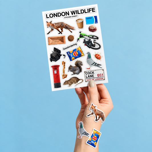 A hand holds a sticker sheet titled 'LONDON WILDLIFE stickers' against a solid light blue background. The sheet contains various animal and London-themed stickers, including a fox, pigeon, squirrel, post box, and a red bus.