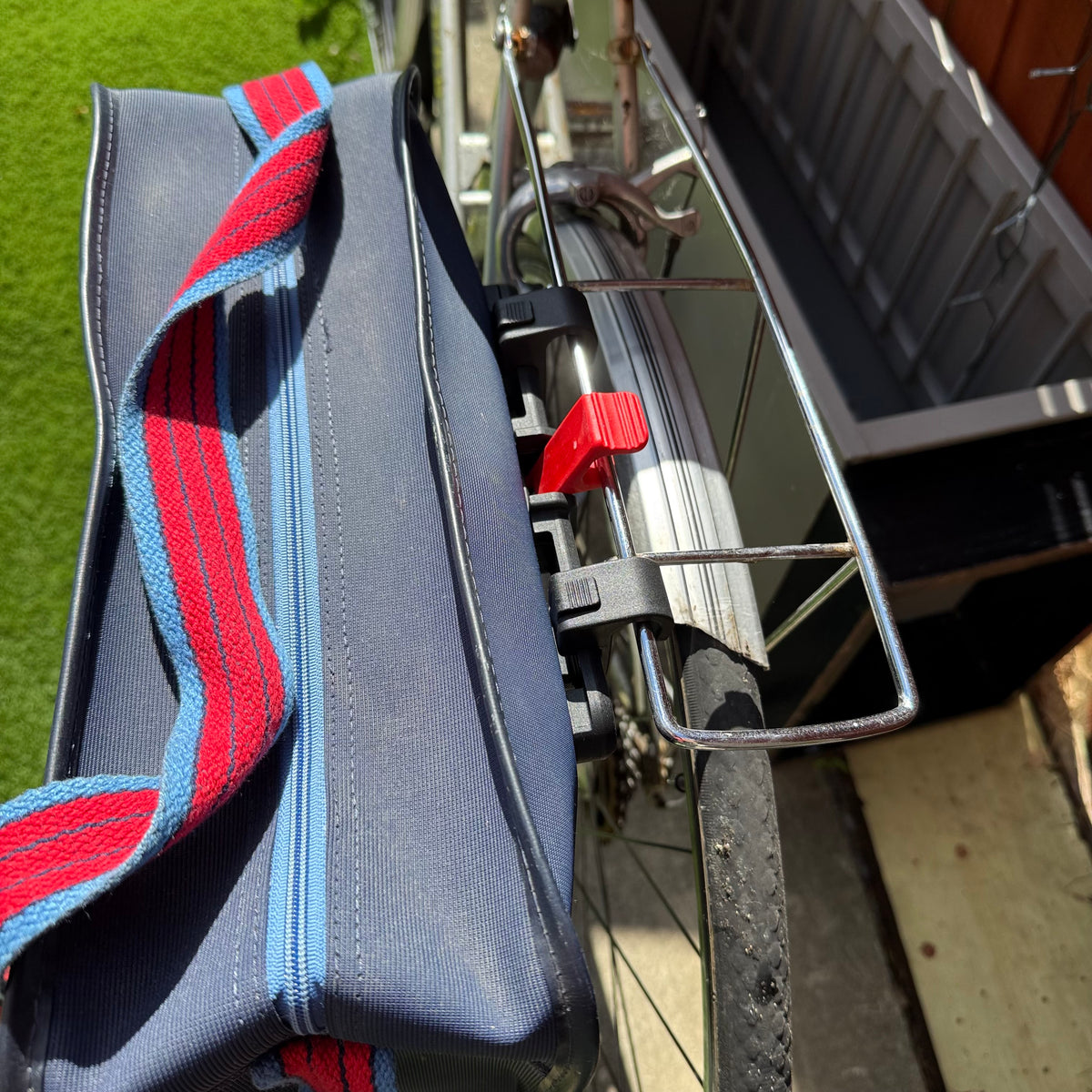 A close-up of a navy blue bicycle pannier bag with red and blue striped webbing straps, attached to a bicycle's rear rack in an outdoor setting.