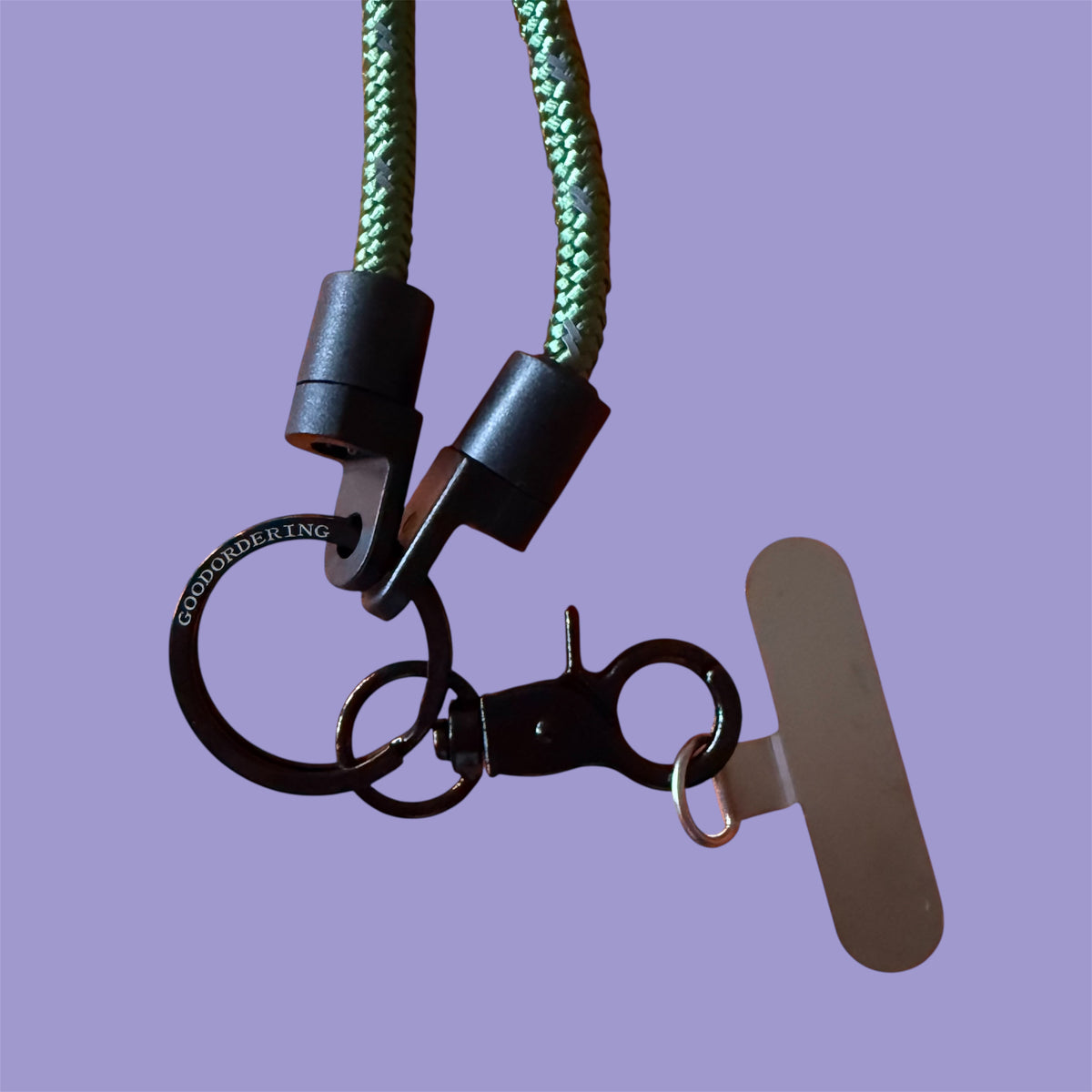 A close-up of a keychain or bag strap accessory with a green braided cord, black plastic and metal hardware, a black ring engraved with 'GOODORDERING', and a flat metallic tag, shown against a solid lavender background.