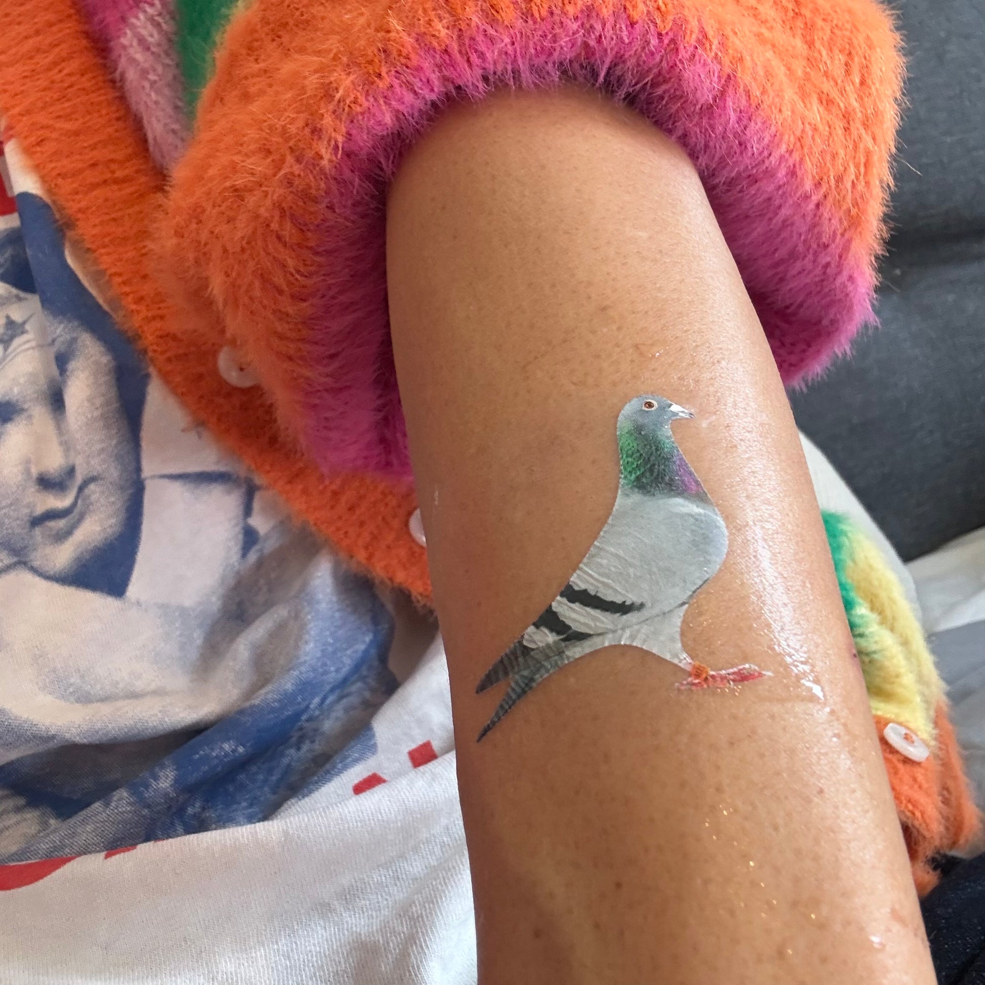 A temporary tattoo of a pigeon applied to a person's forearm, shown next to a colorful fuzzy sleeve.