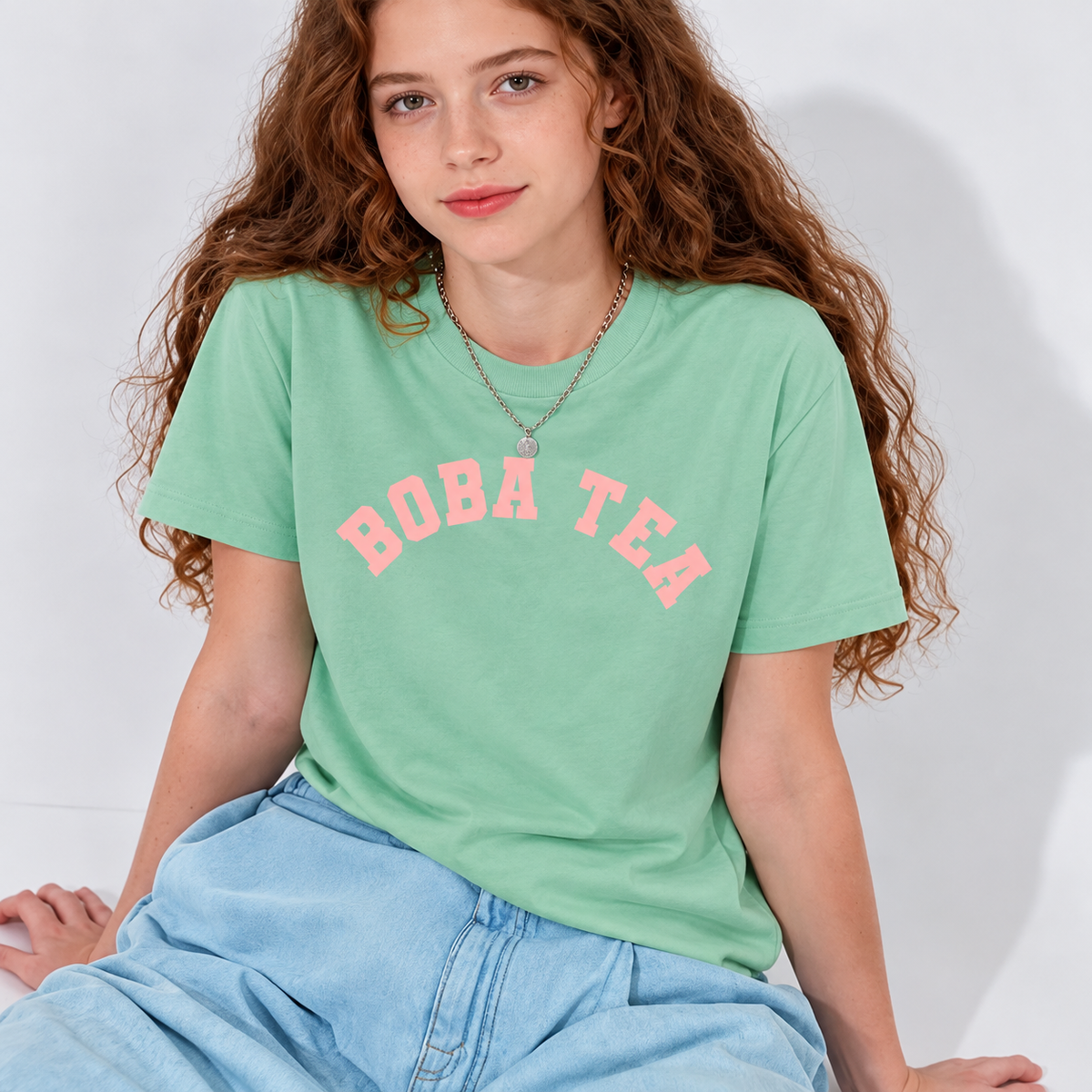 A young woman with long curly brown hair wearing a mint green t-shirt with 'BOBA TEA' printed in pink arched letters across the chest, paired with light blue jeans. Varsity graphic tee inspired by Asian bubble tea culture