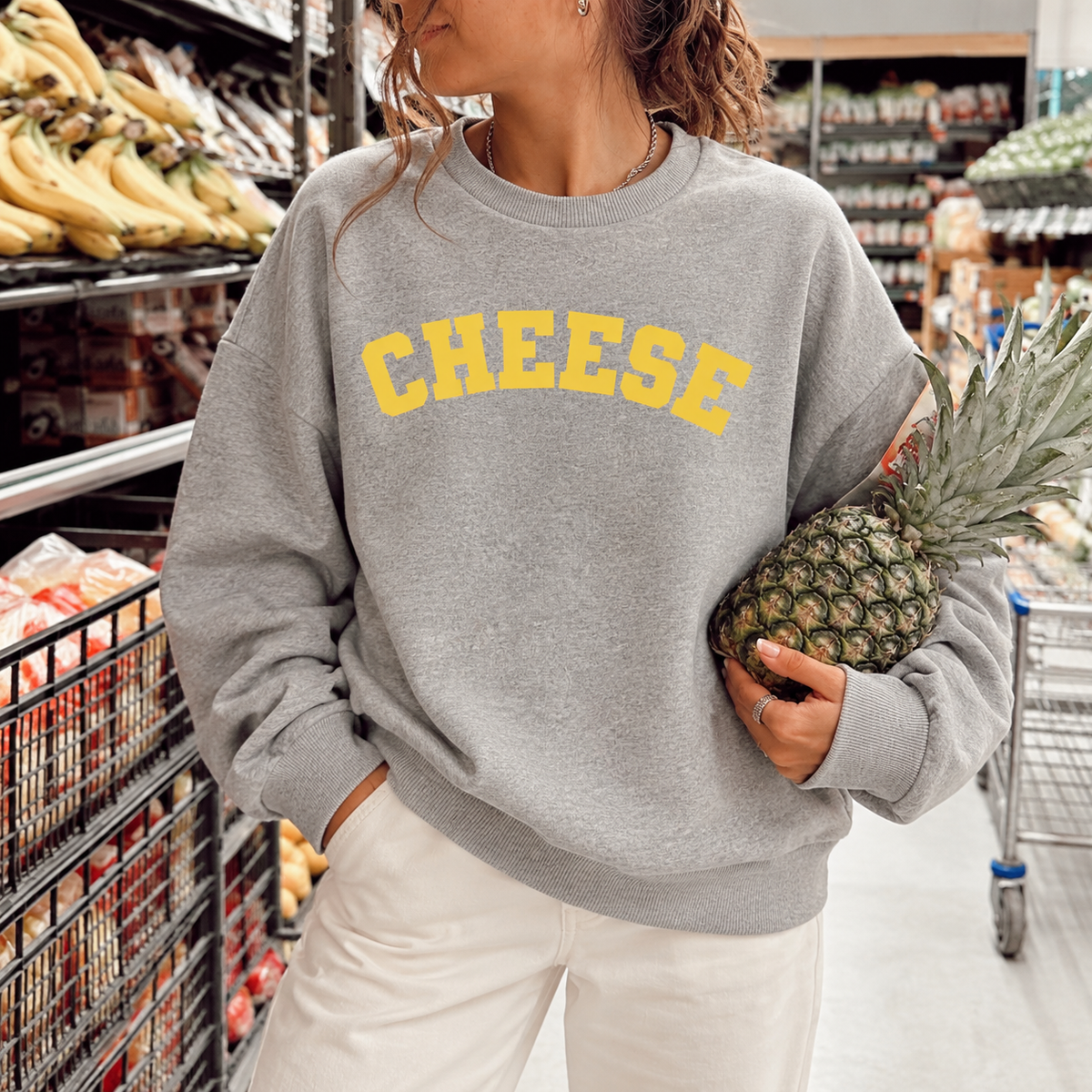 A woman in a grocery store aisle wearing a heather gray sweatshirt with 'CHEESE' printed in yellow across the chest, paired with white pants and holding a pineapple.