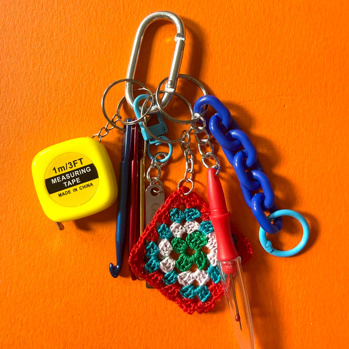 A carabiner keychain holding multiple colorful accessories, including a yellow measuring tape, blue and red crochet hooks, a crocheted granny square charm, a blue chain, and a red pen, set against a bright orange background. colourful-crochet-hooks-craft-keyring