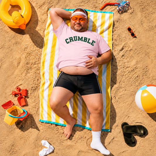 A man lies on a yellow and white striped beach towel on sand, wearing a pink t-shirt with 'CRUMBLE' printed in green, black swim trunks, white socks, and orange-tinted sunglasses. Beach toys and sandals are scattered around him.