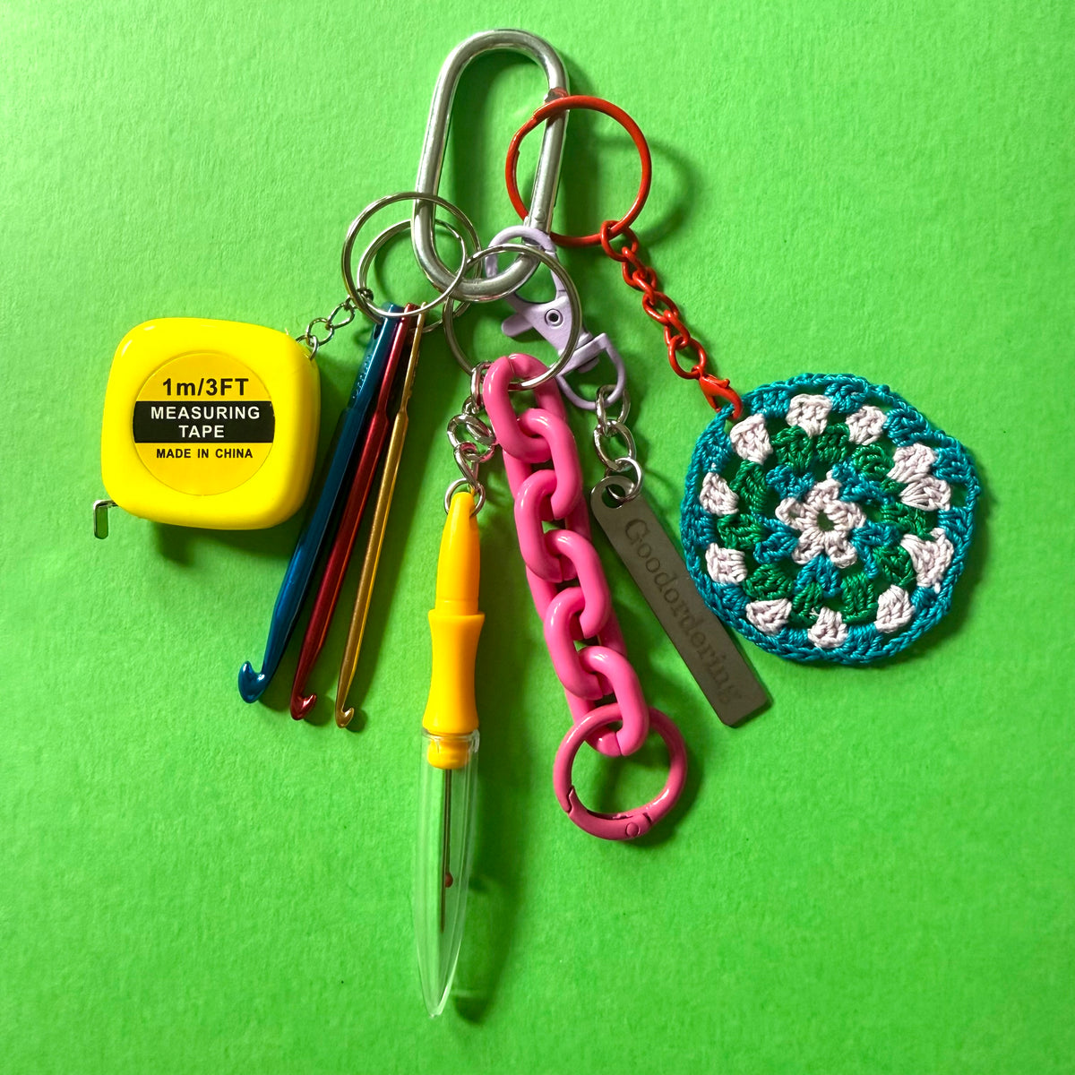 A crochet keychain set featuring a silver carabiner, a pink chain link, a red chain link, a yellow measuring tape, three crochet hooks, a pen, and a crocheted granny square, all arranged on a bright green background. - east-london-handmade-craft-bag-charm