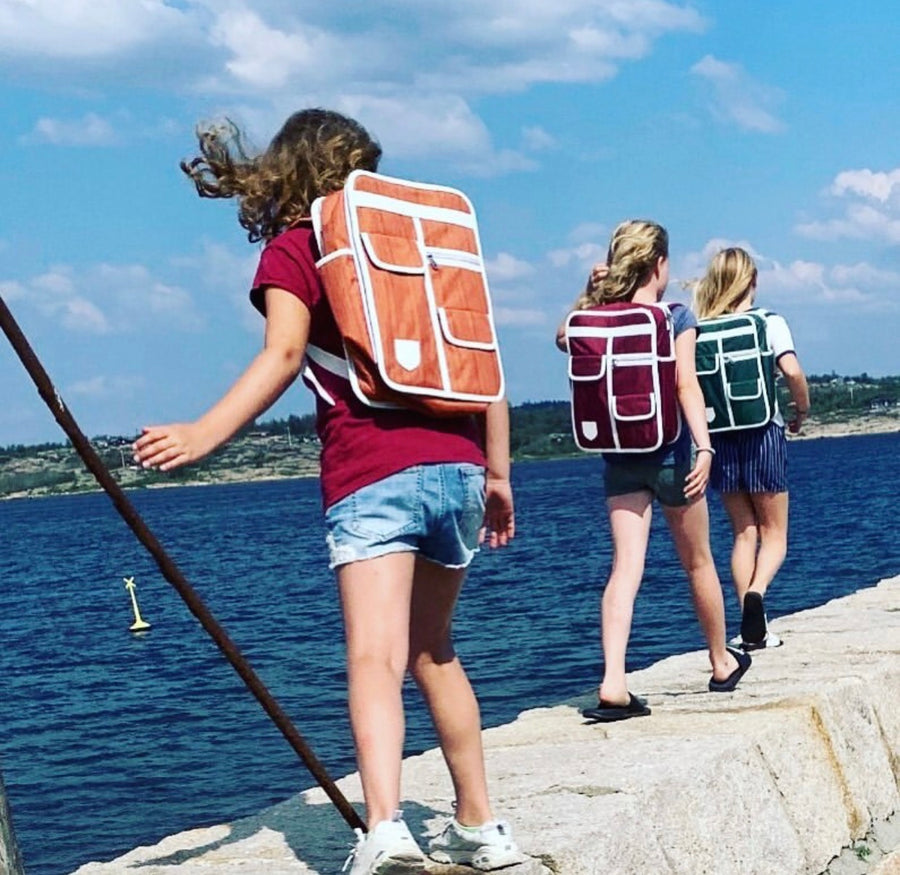 Three girls walking along a stone pier by the ocean, each wearing a different colored goodordering backpack. The girl in the foreground wears an orange backpack, and the background shows blue water and sky.