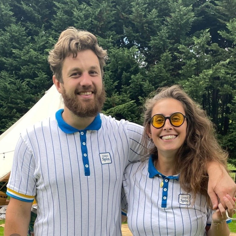 A smiling man and woman standing outdoors in front of trees, wearing matching white polo shirts with thin blue pinstripes, blue collars, and small square logos on the chest and maroon goodordering glasses