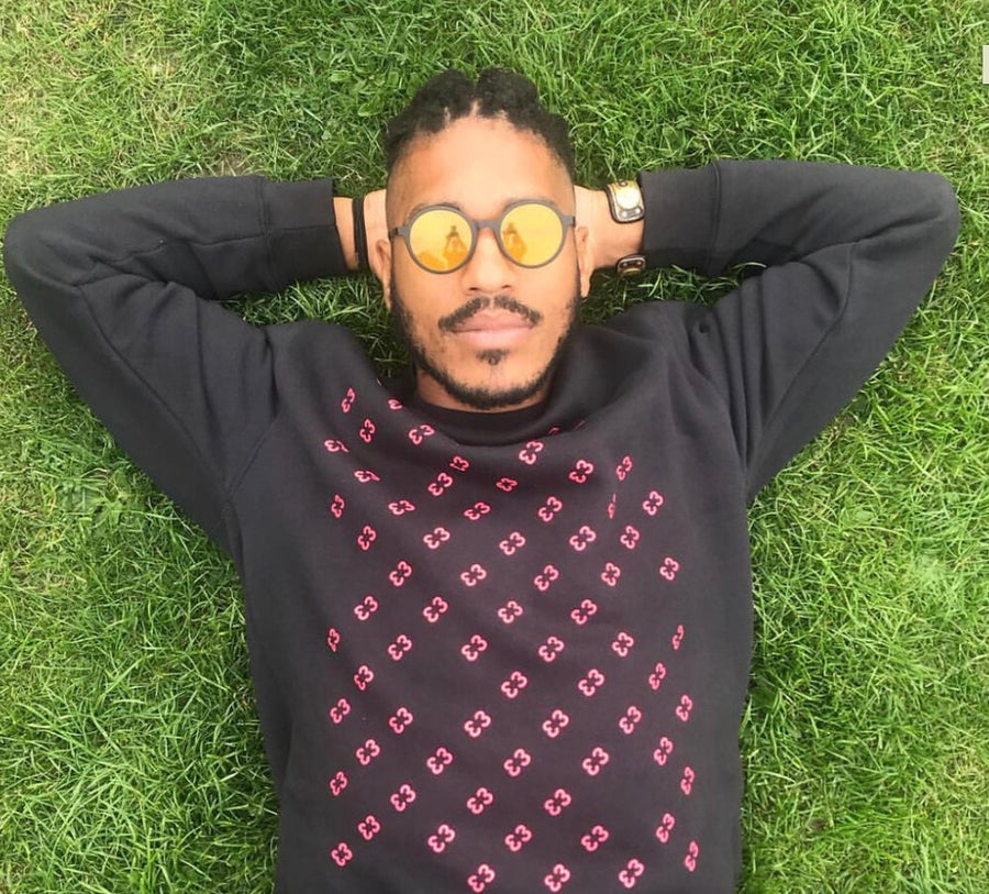A man lying on green grass wearing a black long-sleeve sweatshirt with a pink geometric pattern, round sunglasses with yellow mirrored lenses, and a watch.