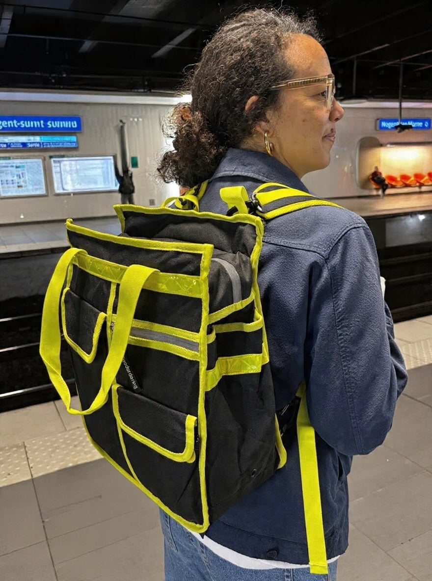 A woman wearing a goodordering black and neon yellow high-visibility utility backpack, shopper bag photographed from behind in what appears to be a train station or transit platform.