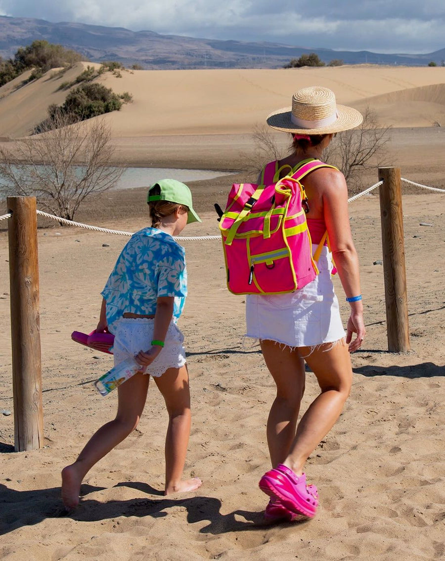 A woman and a child walking on a sandy beach path, viewed from behind. The woman wears a straw hat, a white skirt, and a bright pink backpack, while the child wears a green cap, a blue patterned shirt, and white shorts.