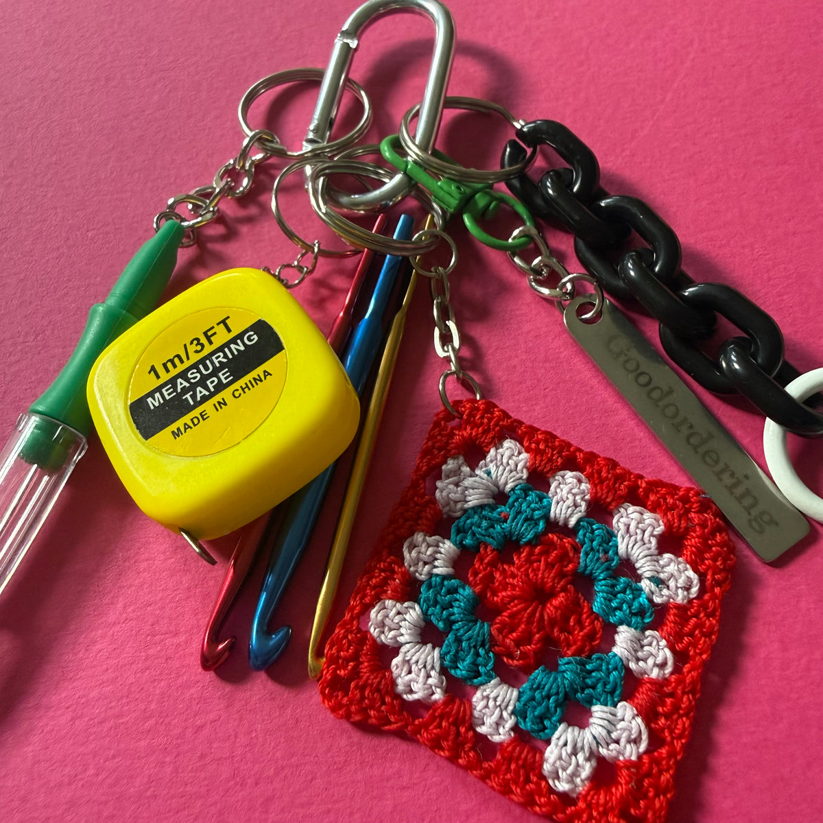 A handmade crochet keychain set on a bright pink background, featuring a red, teal, and white granny square, a black chain, a silver metal tag engraved with 'woodbording', a yellow measuring tape, and crochet hooks. handmade-crochet-granny-square-bag-charm