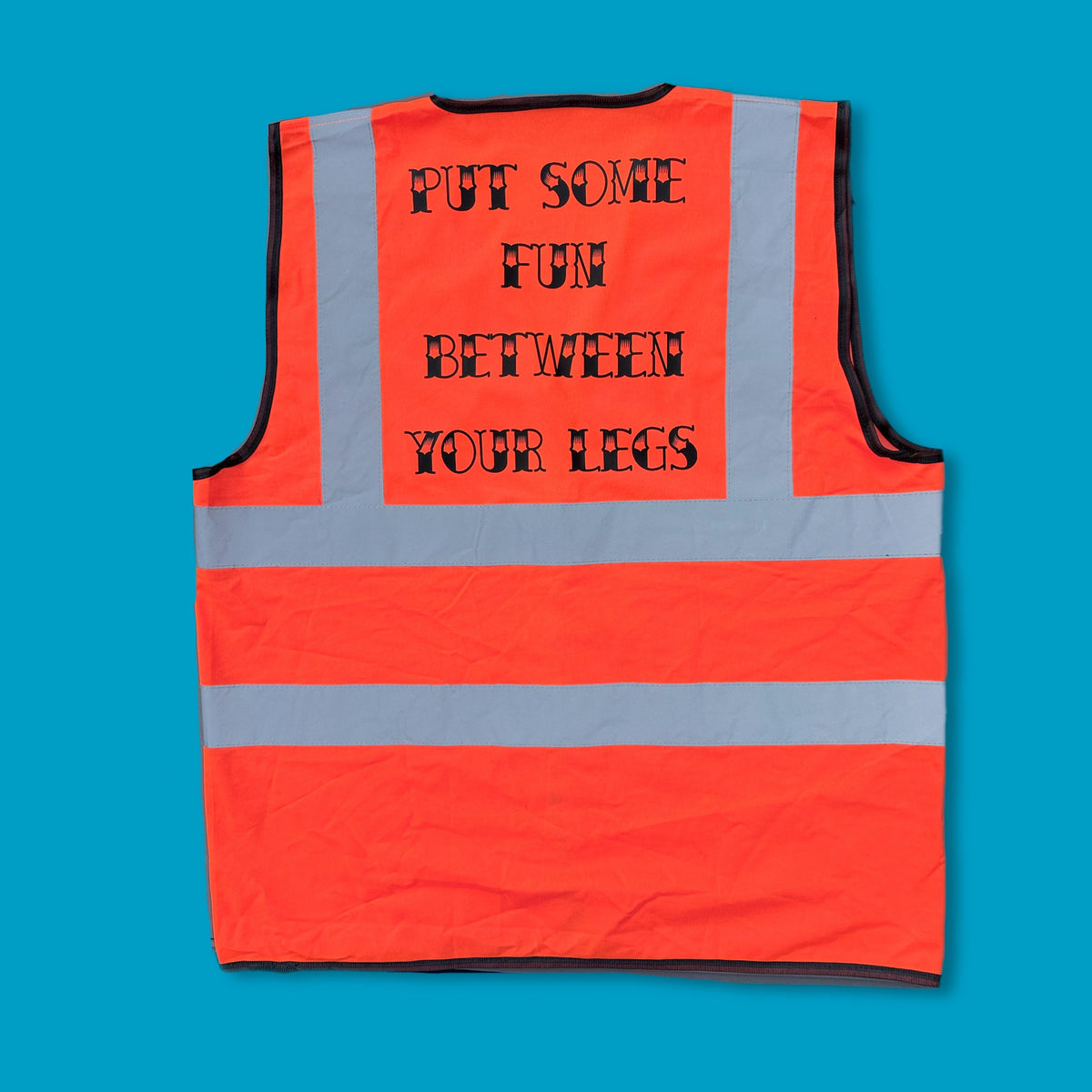 An orange high-visibility safety vest with reflective silver stripes, featuring the humorous text 'PUT SOME FUN BETWEEN YOUR LEGS' printed in black on the back, laid flat against a solid blue background.