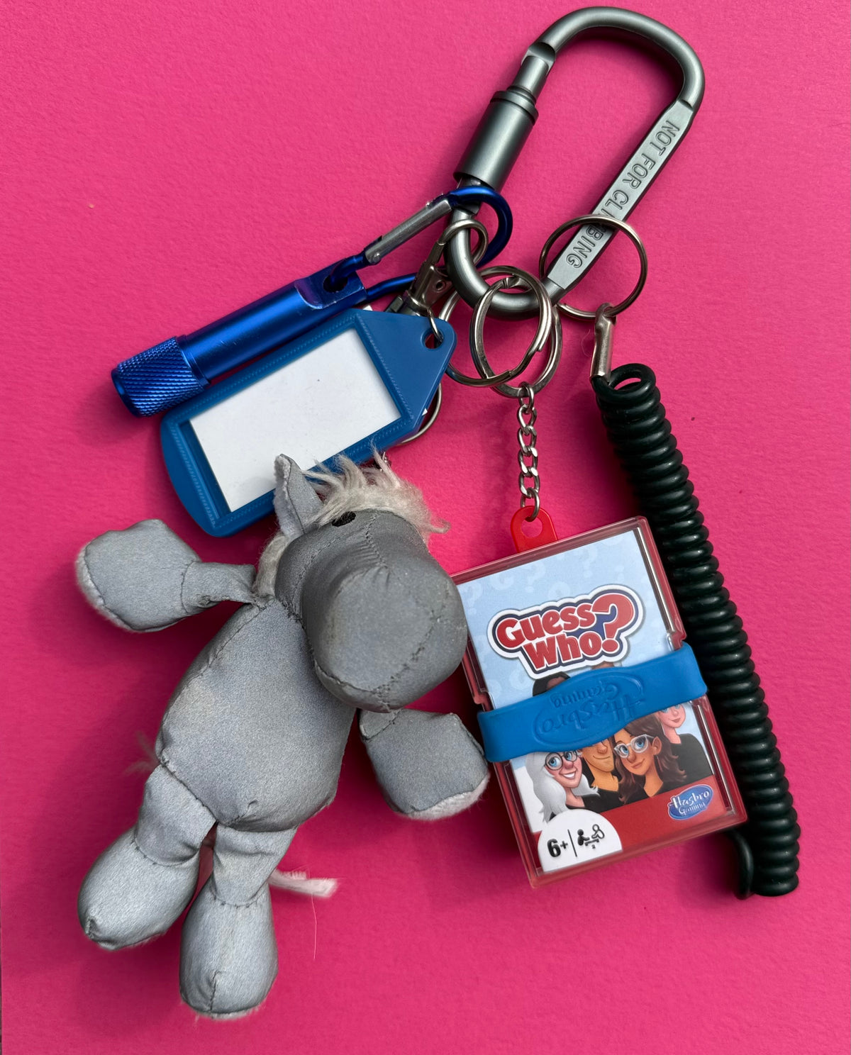 A keychain bundle featuring a gray plush toy, a blue flashlight, a blue ID tag, a black coiled cord, a silver carabiner, and a Guess Who game pack, arranged on a bright pink background.