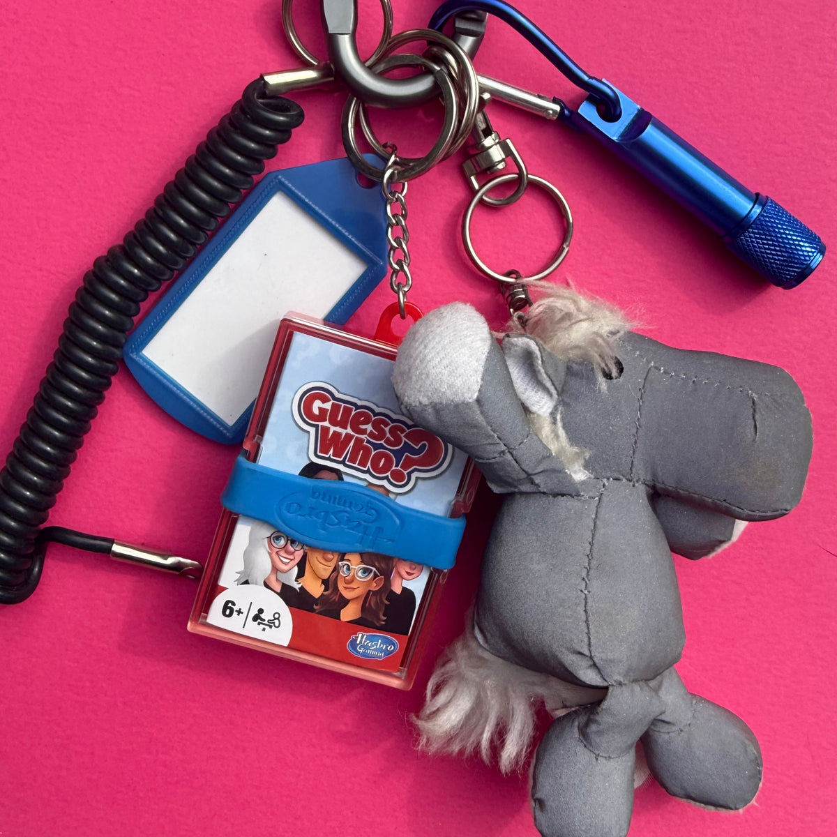 A gray plush elephant keychain with a blue band reading 'Guess Who' and a small Hasbro logo, attached to metal rings with a coiled black cord, blue card holder, and blue flashlight on a bright pink background.