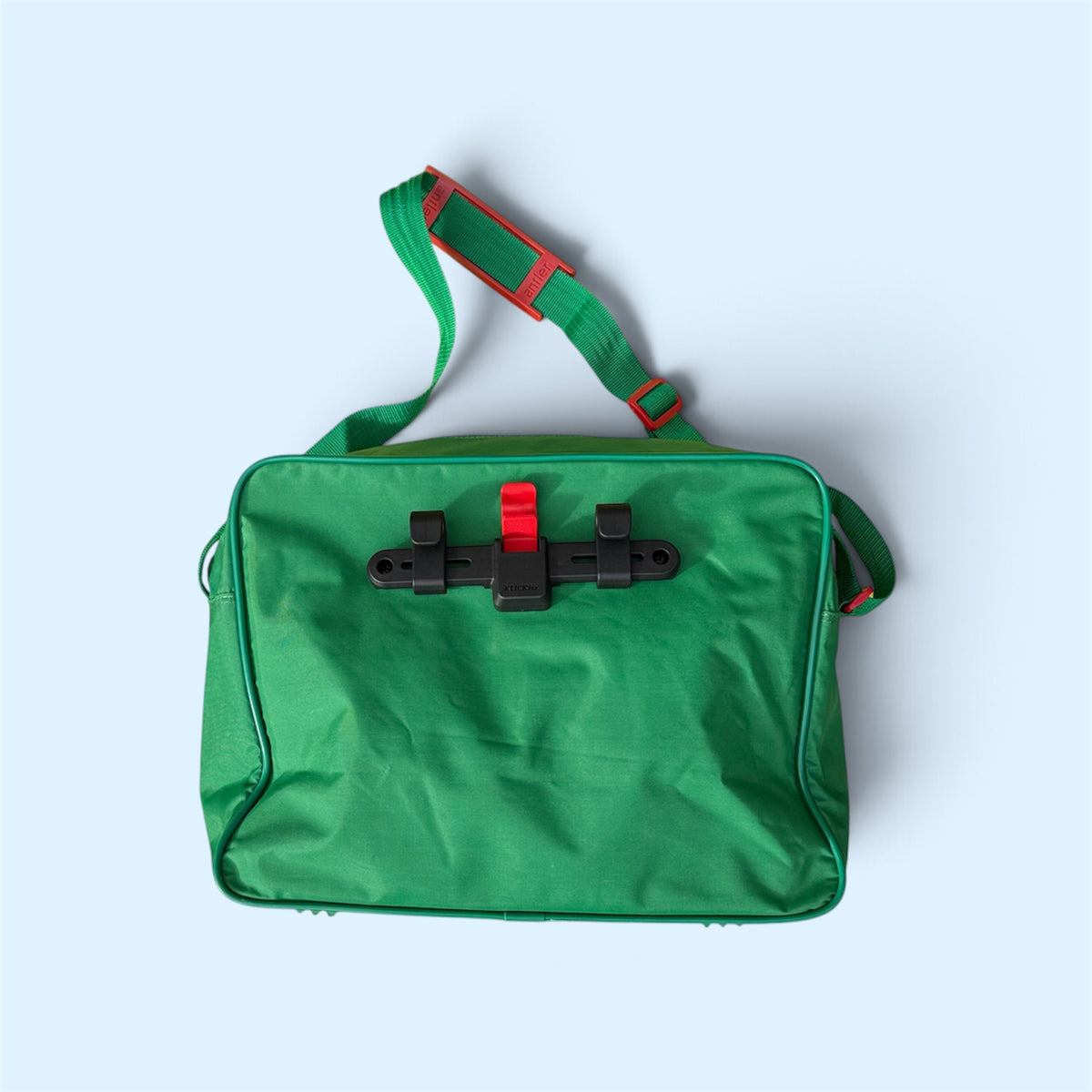 A green rectangular utility bag with a black plastic latch closure and a red release button, featuring a matching green shoulder strap with red accents, set against a light blue background.
