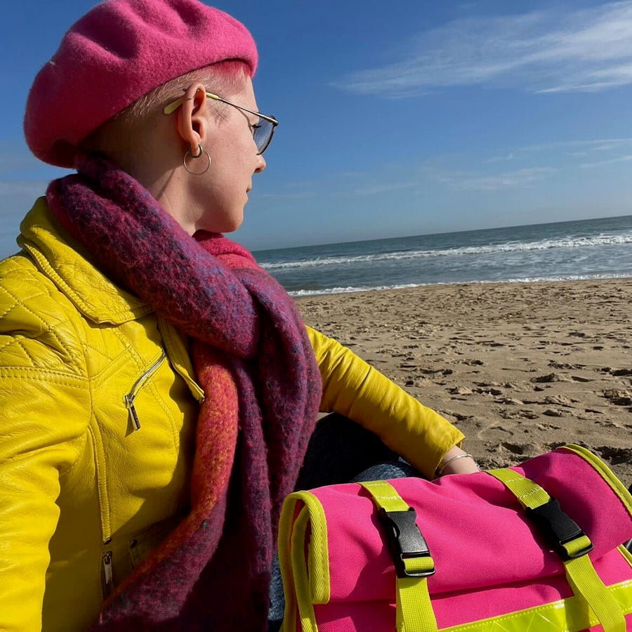 A person sitting on a sandy beach wearing a bright pink beret, a yellow jacket, and a pink and purple scarf, with a matching pink bag in the foreground.
