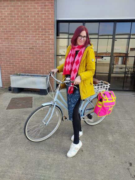 A woman with red hair and glasses stands on a concrete sidewalk next to a light blue bicycle, wearing a bright yellow jacket, a pink and red diamond-patterned scarf, and black leggings. She is holding the bicycle's handlebar, and a pink and yellow bag is attached to the rear of the bicycle.