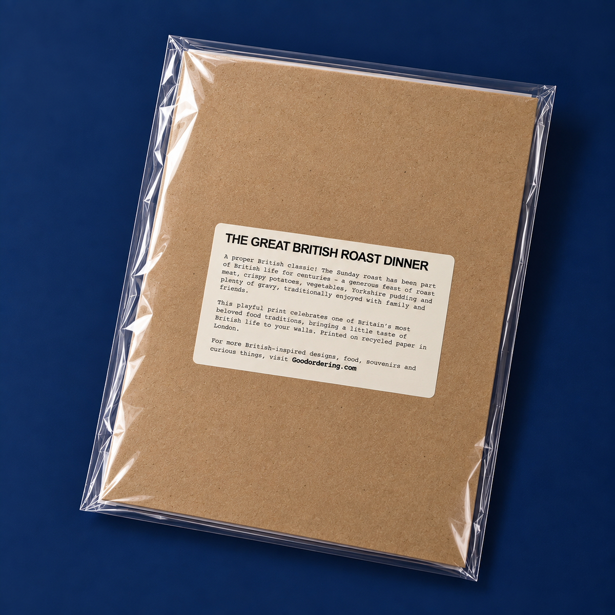 A square kraft paper recipe book or booklet wrapped in clear plastic packaging, shown at an angle on a dark blue background. The front has a white label with black text reading 'THE GREAT BRITISH ROAST DINNER' and a short description.