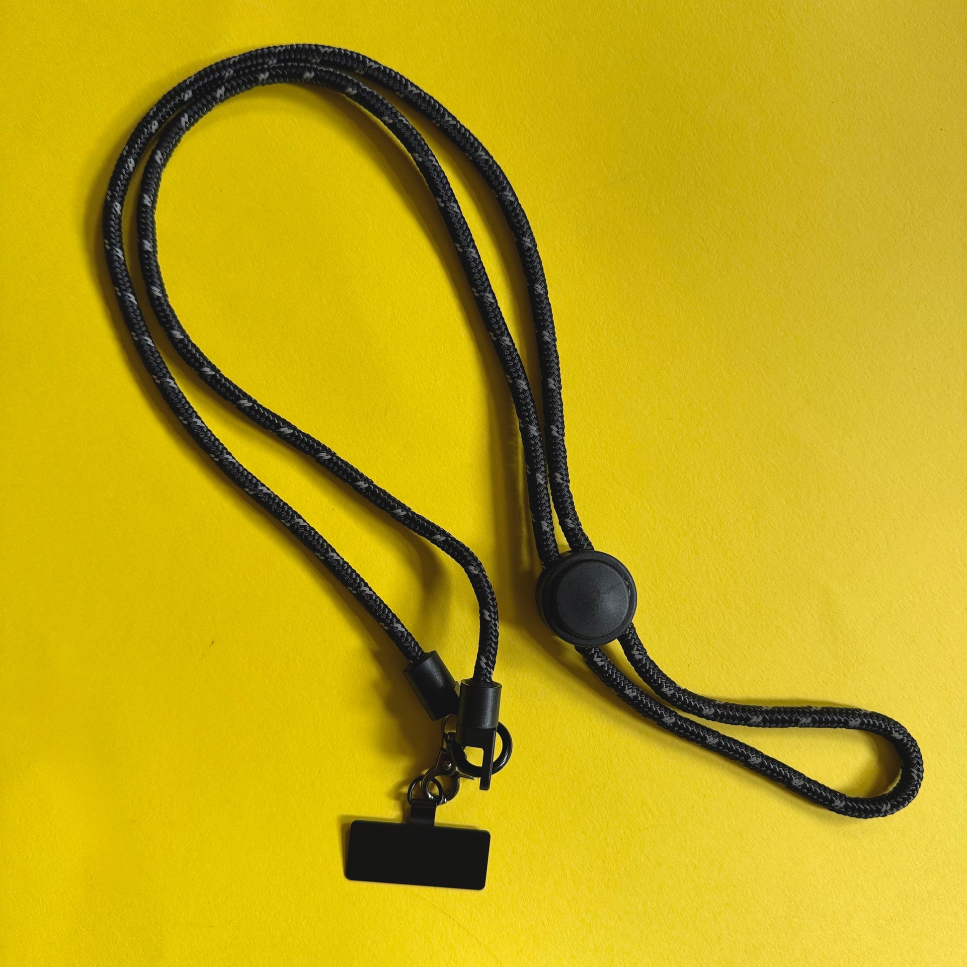 Black braided cord lanyard with a round black plastic toggle, metal clip, and small rectangular black tag, laid on a solid yellow background.