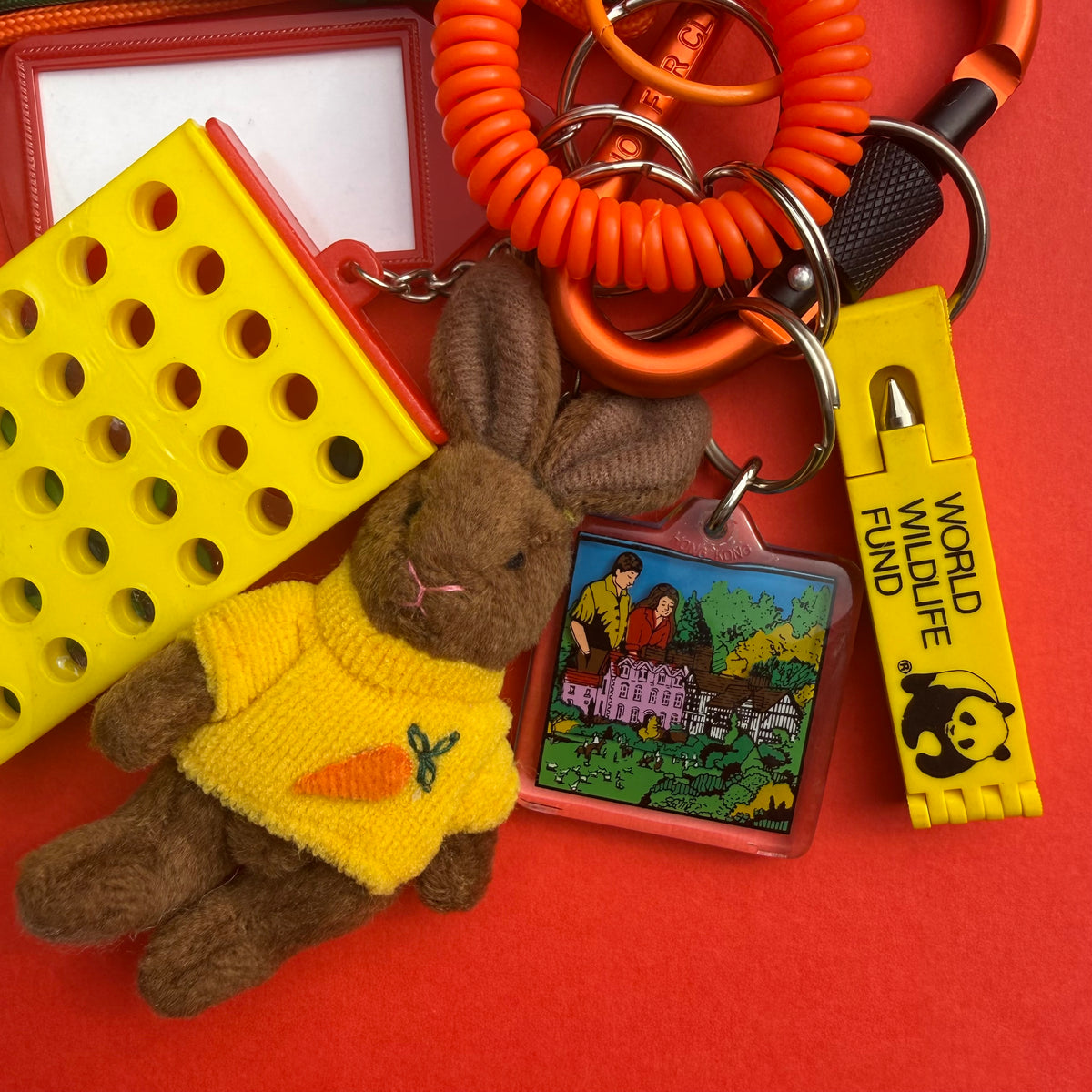 A colorful vintage keychain set featuring a brown plush bunny wearing a yellow sweater with a carrot patch, a yellow rectangular pegboard accessory, an orange coiled cord, a square cartoon charm, and a yellow 'World Wildlife Fund' tag, all arranged on a bright red background.