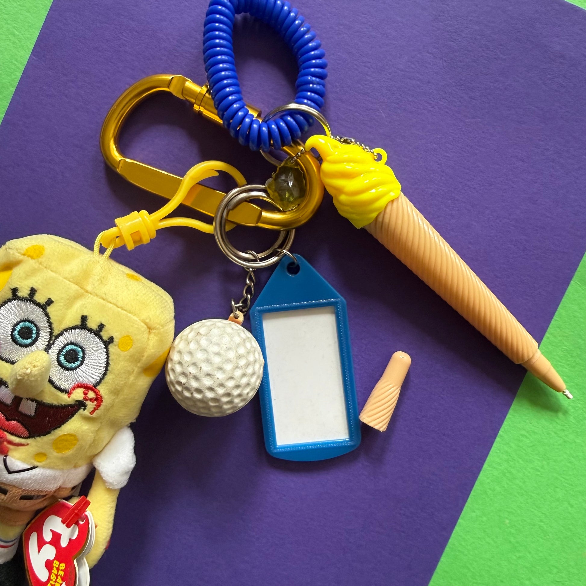 A SpongeBob-themed keychain set laid on a purple and green background, including a yellow plush character, a blue coiled strap, a gold carabiner, a blue tag, a white golf ball charm, a yellow-and-beige novelty pen, and a small beige piece.