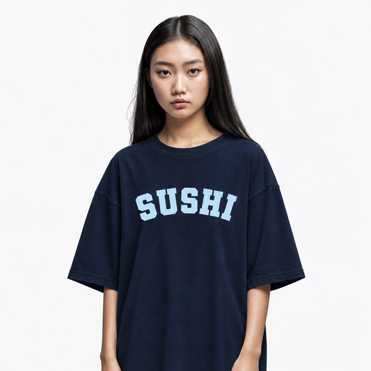 A young woman modeling an oversized dark navy blue t-shirt with the word 'SUSHI' printed across the chest in light blue varsity-style lettering, set against a plain light gray background.