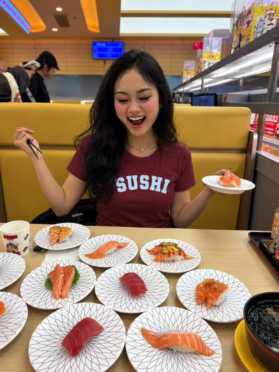 A woman sitting in a sushi restaurant wearing a maroon t-shirt with 'SUSHI' printed in white. She is holding chopsticks and a small plate of sushi, with multiple plates of sushi spread out. sushi varsity tshirt  on the table in front of her.