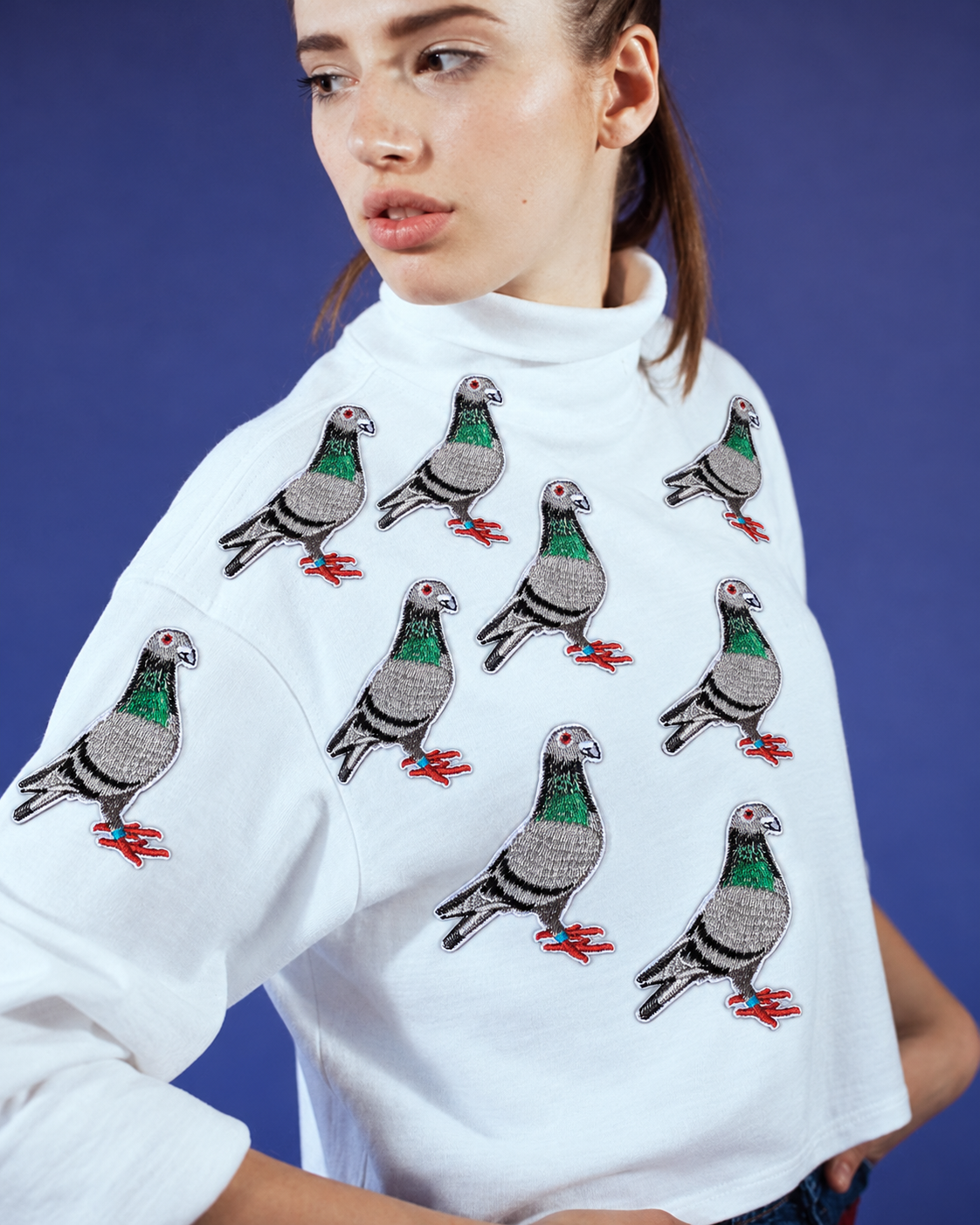 A woman wearing a white turtleneck sweatshirt with embroidered pigeon motifs, posed against a solid blue studio background.