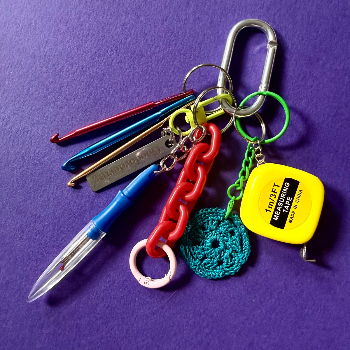 A colorful keychain set featuring a silver carabiner, metal crochet hooks, a blue pen, a red chain link, a green crochet chain, a green circular crochet piece, and a yellow tape measure keychain, arranged on a textured purple background.